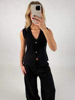 En person i sort vest og bukser holder en mobiltelefon op foran ansigtet, mod en simpel, ensfarvet baggrund.