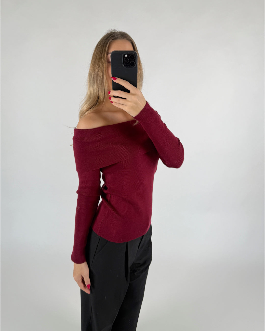 En person tager en selfie med en smartphone, iført en off-shoulder bordeaux trøje og sorte bukser, i et enkelt, lyst rum.