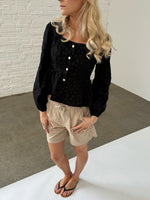 En kvinde med lyst, bølget hår står iført en sort bluse med knapper og beige shorts. Hun har sorte klipklappere på en grå beton- og hvid baggrund.