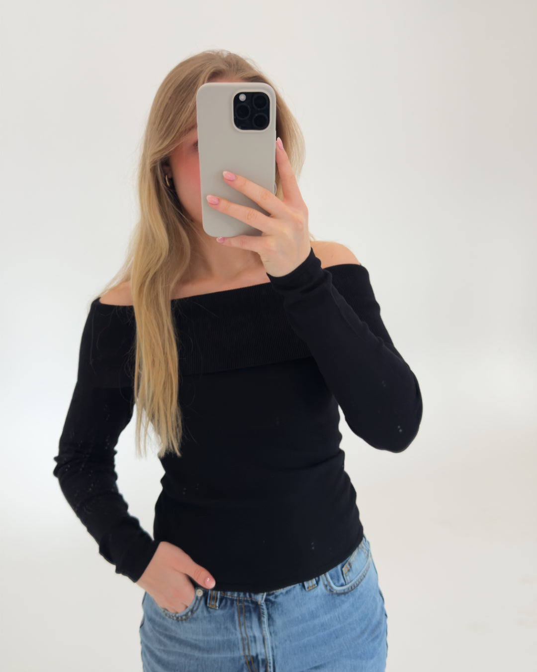 En person holder en telefon op foran ansigtet, iført en sort off-shoulder trøje og blå jeans, stående i et hvidt, neutralt rum.