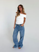 En person står afslappet iført en hvid top og blå jeans i et lyst, minimalistisk rum. Håret er langt og bølget, og hænderne er i lommen.