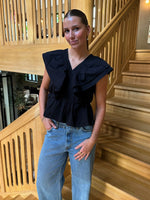 En person står afslappet med hænderne i lommerne, iført en sort bluse og jeans. Baggrunden er en indendørs trætrappe med moderne design.