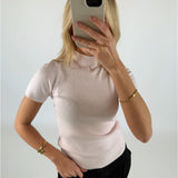 En person iført en lys t-shirt holder en smartphone foran ansigtet, i et hvidt, minimalistisk rum. Personen bærer guldsmykker på begge hænder.