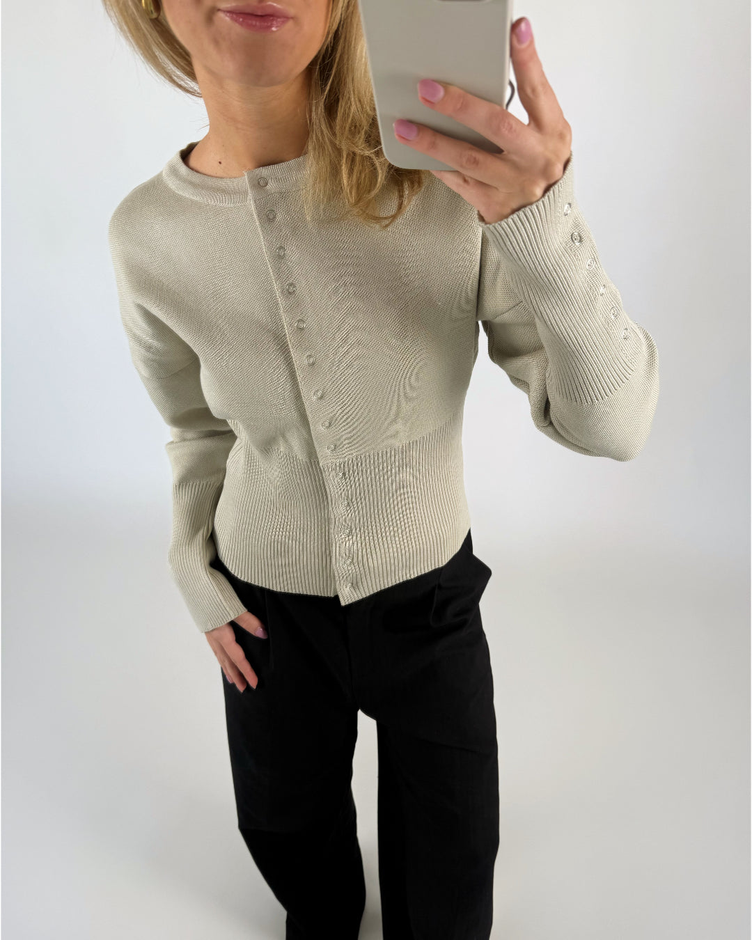 En person iført en beige sweater med knapper tager en selfie. Personen bærer sorte bukser, og baggrunden er en neutral hvid farve.