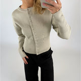 En person iført en beige sweater med knapper tager en selfie. Personen bærer sorte bukser, og baggrunden er en neutral hvid farve.