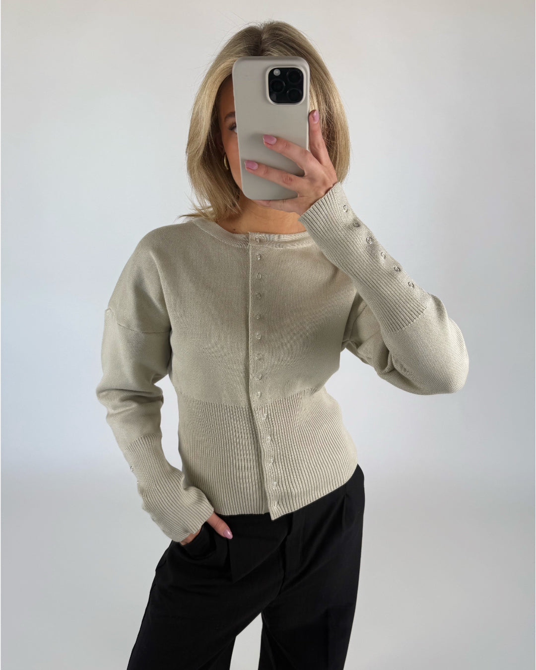 En person i en lys beige cardigan holder en smartphone op foran ansigtet. Personen står i et rum med en hvid baggrund og har en hånd i lommen på sine sorte bukser.