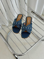 Sorte denim-sandaler med nitter ligger stille på en moderne metalstol. Tekst på indersålen: "stephan shoes." Gardiner i baggrunden skaber en rolig atmosfære.