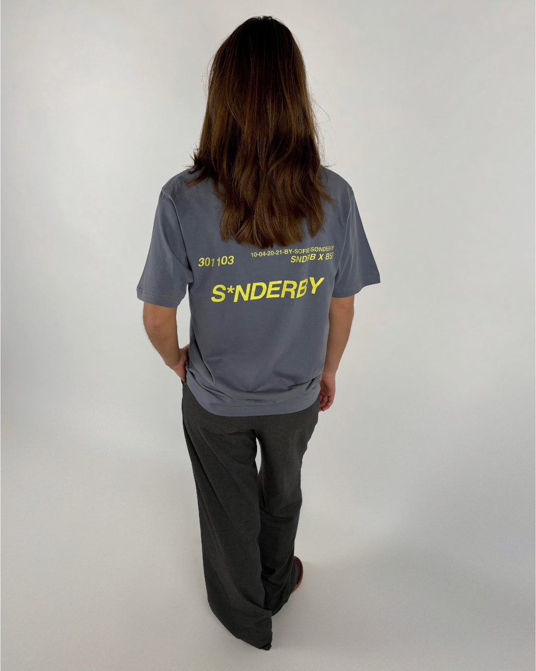 En person står med ryggen til iført en grå T-shirt med gul tekst: "301103 10-04-20-21 BY-SOFFE SØNDERBY SNDB X BS S*NDERBY" og sorte bukser i en lys baggrund.