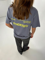 En person med langt hår står i et hvidt rum, iført en grå T-shirt med gul tekst: "30 / 103, 16-04-20-21-BY-SOFIE-SØNDERBY, SNDRB X BSS, S*NDERBY".