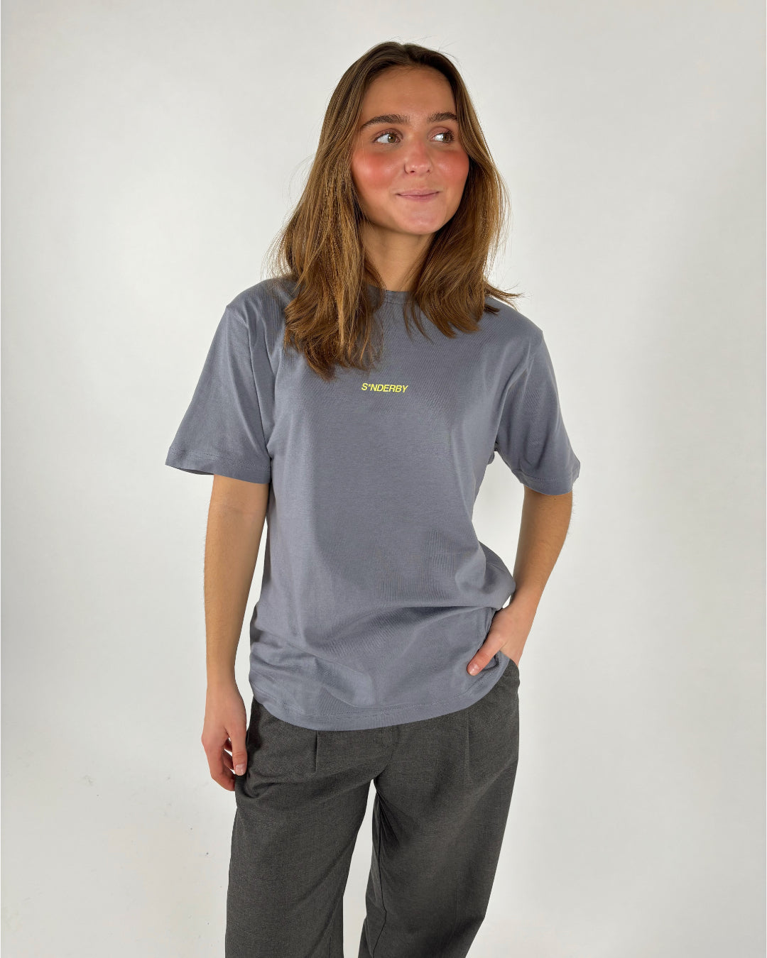 En person i en grå T-shirt med ordene "SØNDERBY" står med hånden i lommen og kigger til siden. Baggrunden er ensfarvet lys.