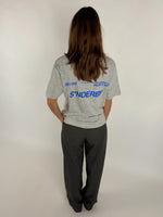 En person med langt hår bærer en grå T-shirt med blå tekst "SØNDERBY 301103" og yderligere tekst i et studie med hvid baggrund.