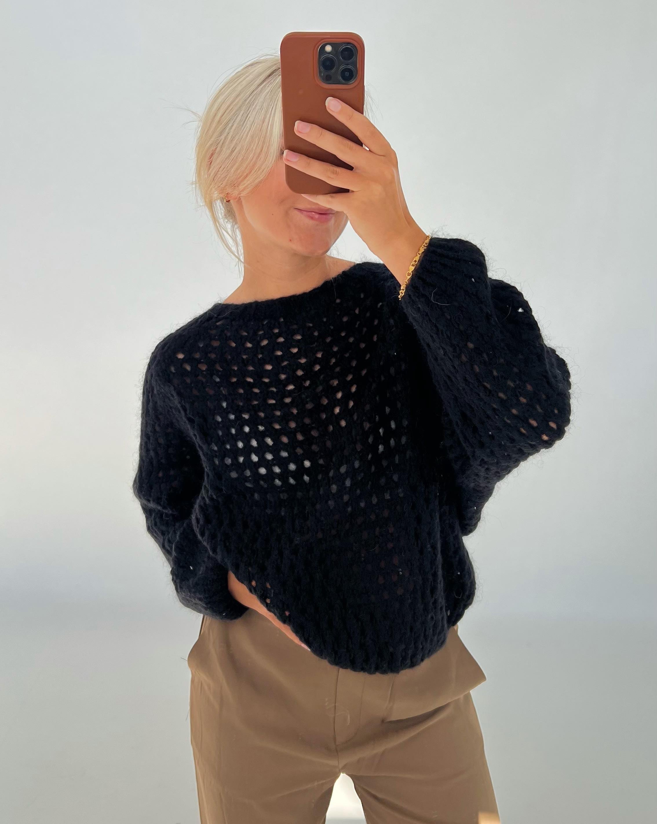 En person iført en sort, hullet sweater tager en selfie med en mobiltelefon i en lys baggrund. Personen har lyst hår og afslappede brune bukser.