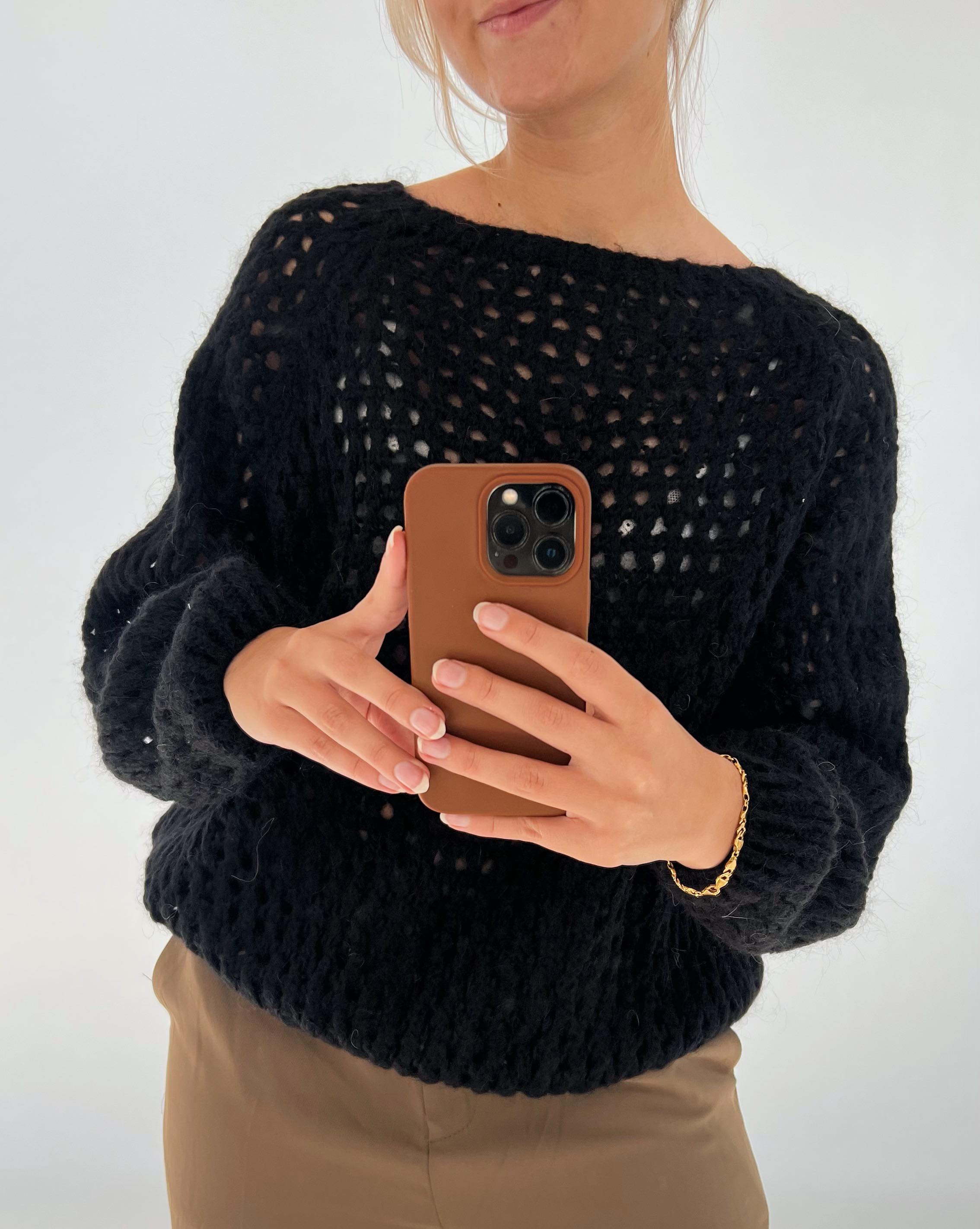 Person tager et spejlbillede med en smartphone. Personen bærer en sort strikket sweater og en brun bukser, med et enkelt guldsmykke om håndleddet. Baggrunden er enkel og lys.