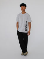 Person står iført en grå t-shirt med teksten "SØNDERBY", sorte bukser og beige sko mod en neutral baggrund.