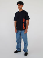 En mand står i afslappet tøj; han bærer en sort t-shirt med ordet "SØNDERBY" skrevet lodret i orange. Han er i et minimalistisk miljø uden distraktioner.