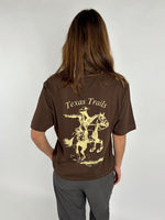 En person iført en brun T-shirt med påtrykt motiv af en cowboy på en hest med teksten "Texas Trails". Personen står mod en neutral, lys baggrund.