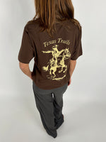 En person med langt hår bærer en brun t-shirt med teksten "Texas Trails" og en illustration af en cowboy på hesteryg i en lys nuance står i et neutralt rum.