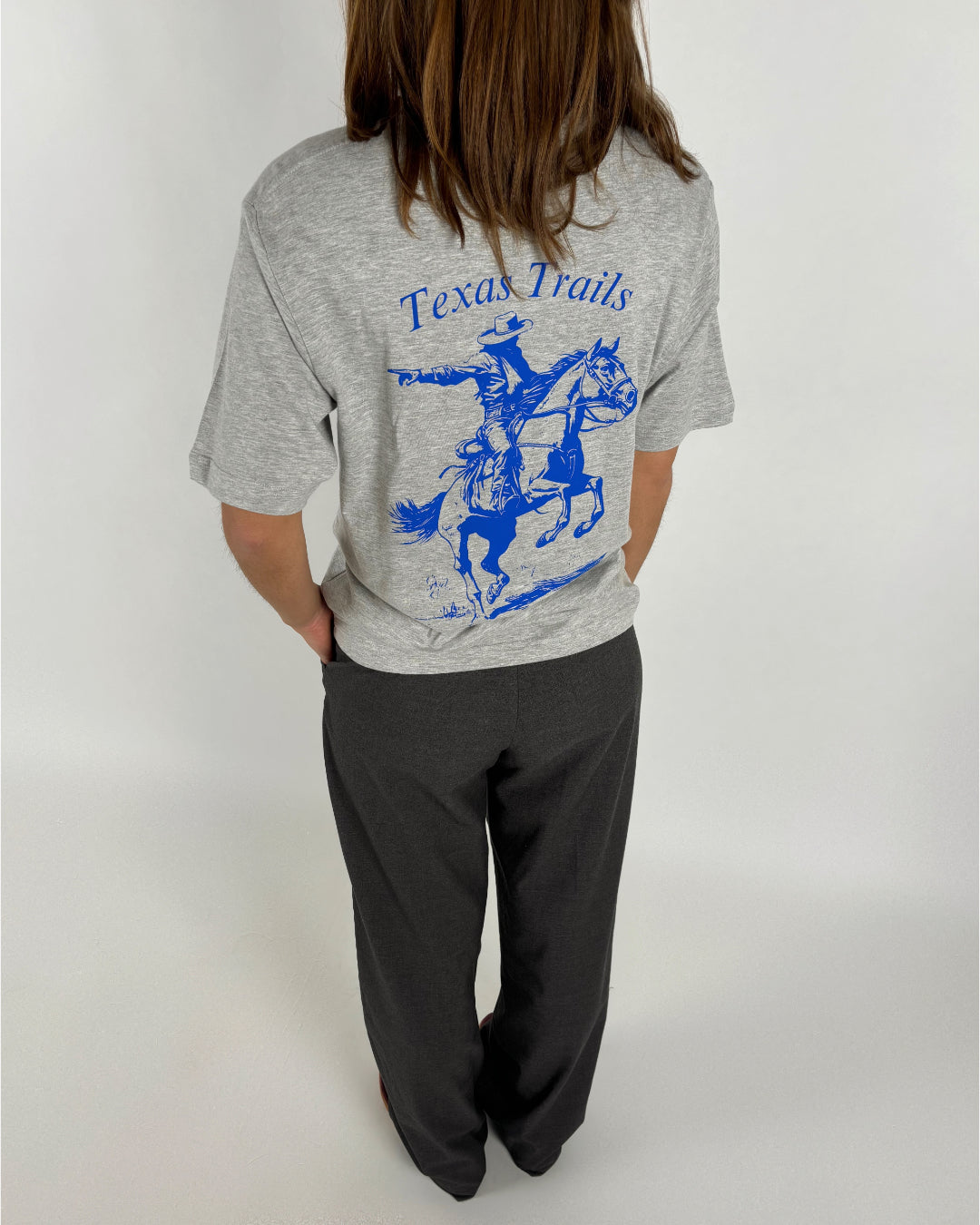 En bagfra-vendt person iført en grå T-shirt med blå grafik af en cowboy på hest og teksten "Texas Trails," står i et neutralt rum med hænderne i bukselommerne.