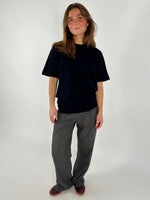 En person står iført en sort t-shirt og grå bukser, smilende let i et neutralt, lyst studieomgivelser. Hun har løst hår og brune sko.