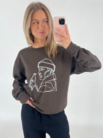 En person iført en brun trøje med hvid stregtegning tager en selfie med en mobiltelefon. Personen står foran en hvid baggrund.