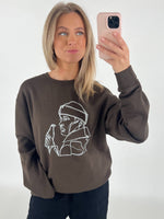 Kvinde står foran et hvidt bagtæppe og tager en selfie med sin telefon. Hun bærer en brun sweatshirt med en hvid minimalistisk tegning af en person, der holder en flaske.