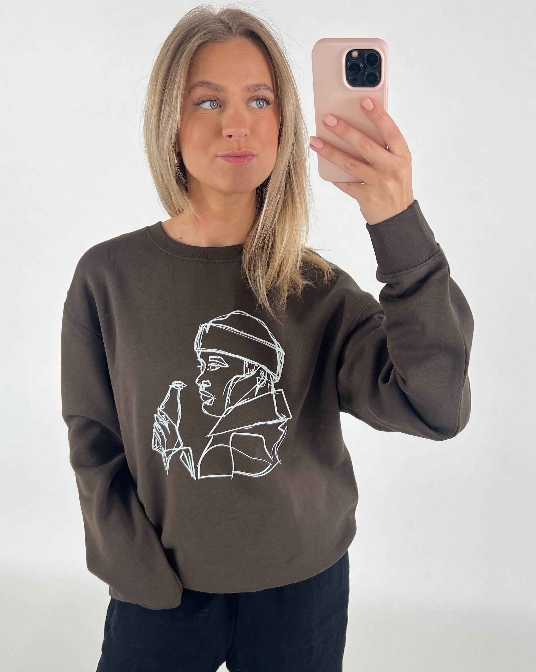 Kvinde står foran et hvidt bagtæppe og tager en selfie med sin telefon. Hun bærer en brun sweatshirt med en hvid minimalistisk tegning af en person, der holder en flaske.
