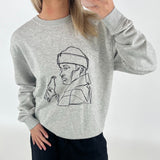 Person iført grå sweatshirt med lineart-tegning af en person med flaske. Personen holder en mobiltelefon foran et neutralt bagtæppe.