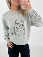 Person iført grå sweatshirt med lineart-tegning af en person med flaske. Personen holder en mobiltelefon foran et neutralt bagtæppe.