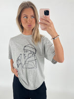 En kvinde fotograferer sig selv med en smartphone, iført en grå T-shirt med en lineart-tegning af en person, der holder en flaske. Hun står i et lyst rum.