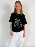 En person iført en sort t-shirt med hvidt lineart-motiv og hvide bukser står i et lyst rum. Vedkommende har langt hår og bærer smykker.