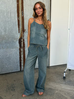 En kvinde i denim jumpsuit står afslappet med hænderne i lommerne. Hun smiler, omgivet af industrielle, rustikke vægge og betongulv.