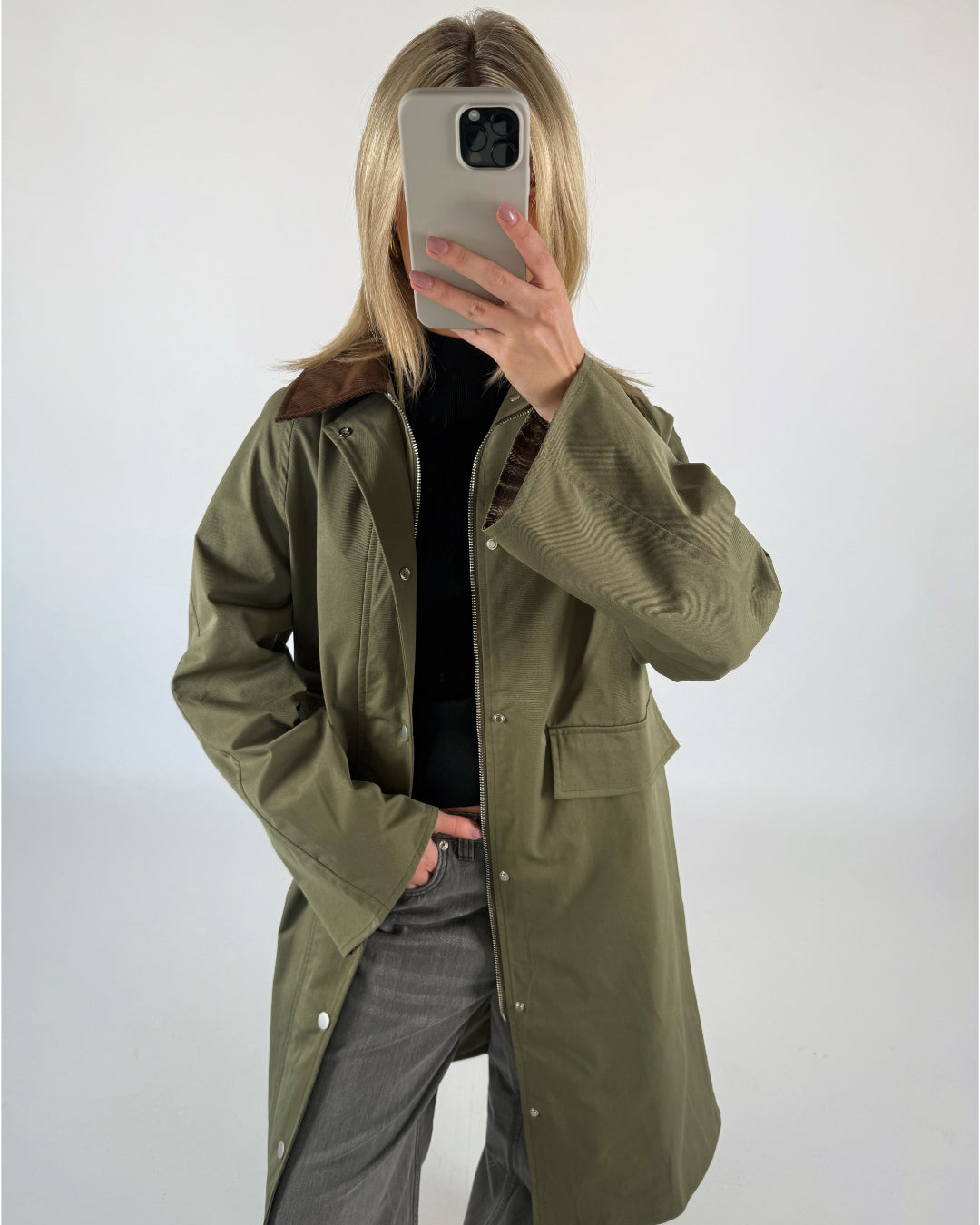 En person iført en grøn frakke tager et spejlfoto med en smartphone i lysegråt etui, i et minimalistisk rum med hvid baggrund.