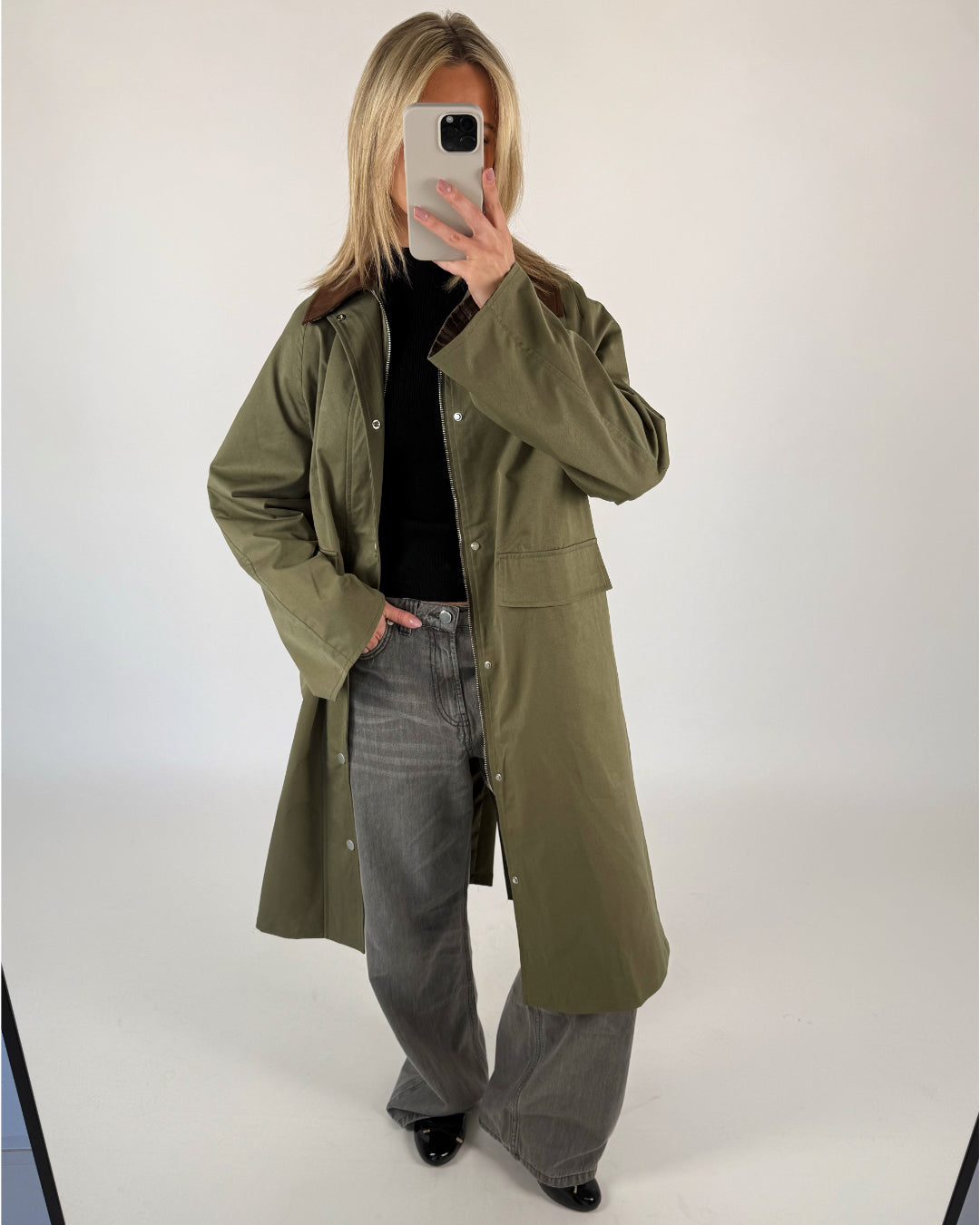 En person iført lang grøn frakke og grå jeans tager et billede med en smartphone i et minimalistisk, lysegråt rum.