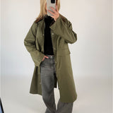 En person iført lang grøn frakke og grå jeans tager et billede med en smartphone i et minimalistisk, lysegråt rum.