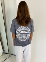 En person står med ryggen til iført en grå T-shirt med teksten "WORLDWIDE" og et globusdesign. Personen er i et minimalistisk rum med et spejl til venstre.