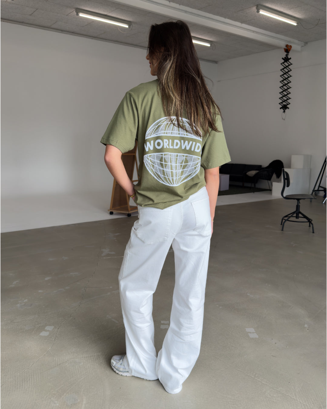 En person iført en olivengrøn T-shirt med teksten "WORLDWIDE" og en globusdesign står i et minimalistisk rum med beton- og hvidmalede overflader, omgivet af møbler.