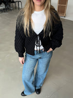En person står iført en sort strikket cardigan og blå jeans, hviler en hånd i lommen, i et minimalistisk kontorrum med lysegråt gulv og stole i baggrunden.