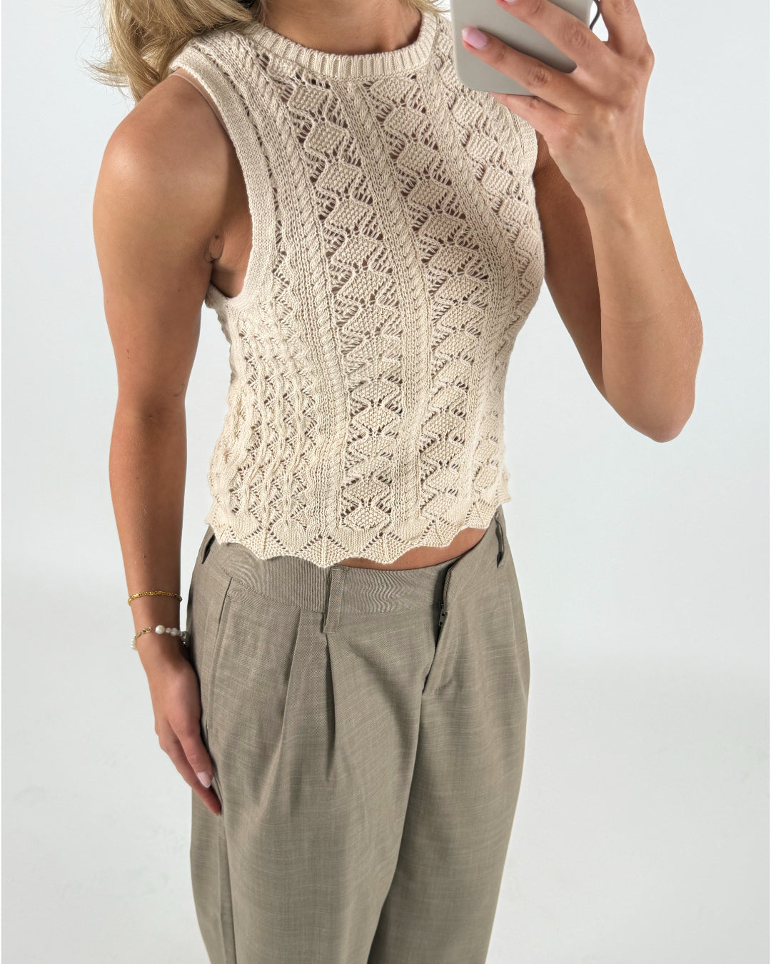 En person iført en ærmeløs, mønsterstrikket beige top holder en telefon, iført lys beige bukser med en perlearmbånd i en minimalistisk, lys baggrund.