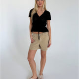 En kvinde iført sort bluse og beige shorts står i et minimalistisk, hvidt rum. Hun har lange, blondehår og sandaler med sløjfe, og poserer afslappet.