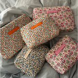 Fem quiltede makeup-tasker med blomsterprint, hver med et "LA PETITE CACHOTIÈRE" -label, ligger oven på et lyseblåt sengetæppe.
