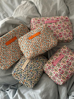 Fem quiltede makeup-tasker med blomsterprint, hver med et "LA PETITE CACHOTIÈRE" -label, ligger oven på et lyseblåt sengetæppe.