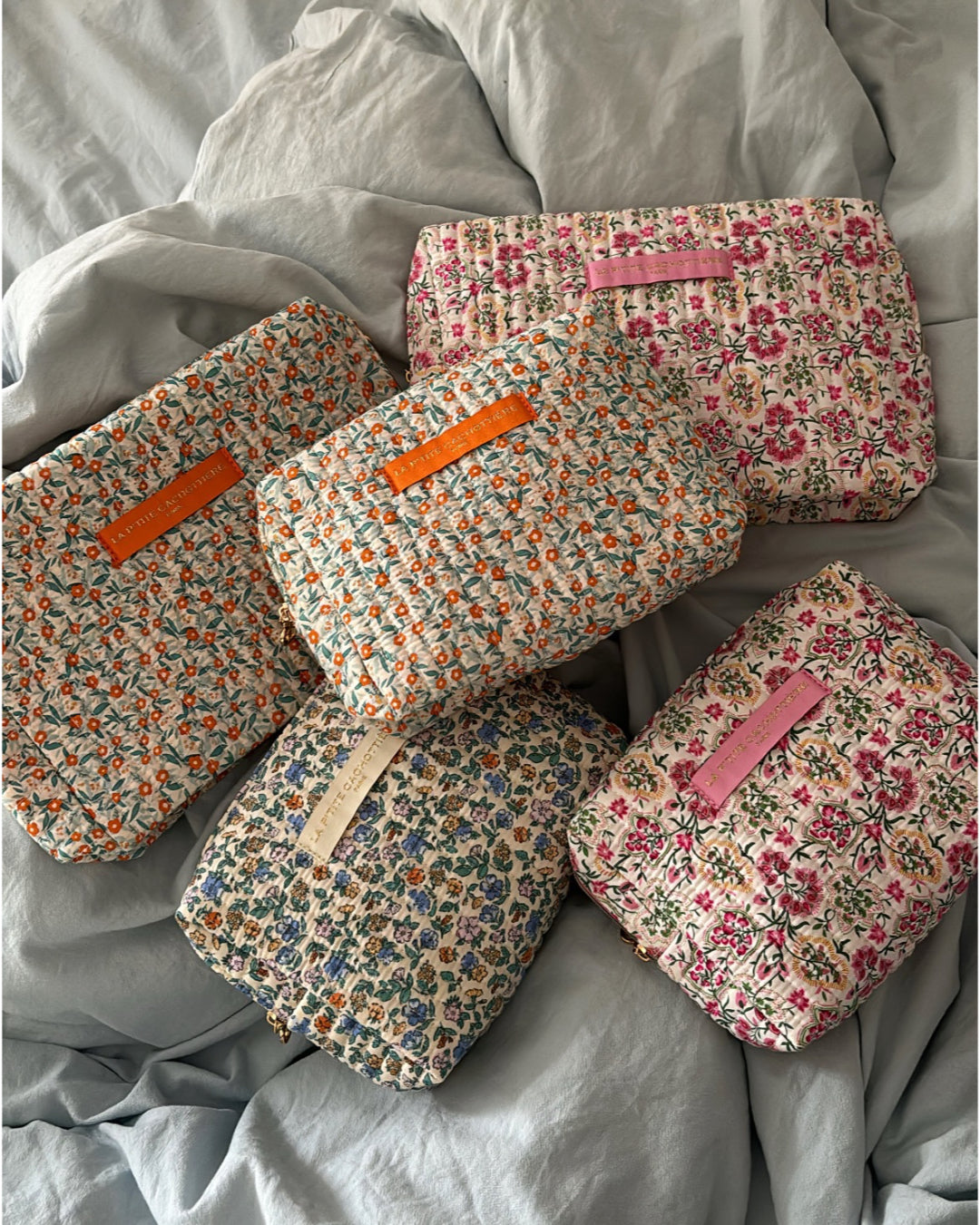 Fem blomstrede toilettasker med mærket "LA PETITE CACHOTIERE" ligger på et lyseblåt dynebetræk. Taskerne har forskellige farver blomster, og mærkerne har orange, lyserød eller beige baggrund.
