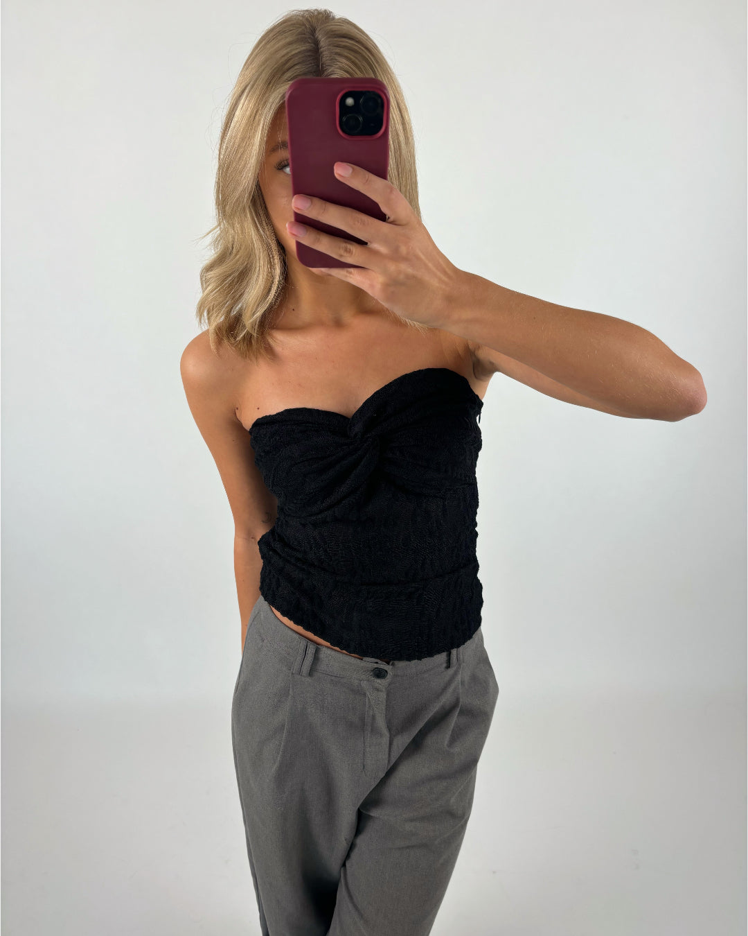 En kvinde tager en selfie med en bordeauxfarvet telefon. Hun er iført en sort top og grå bukser foran en hvid baggrund.