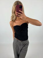 En kvinde tager en selfie med en bordeauxfarvet telefon. Hun er iført en sort top og grå bukser foran en hvid baggrund.