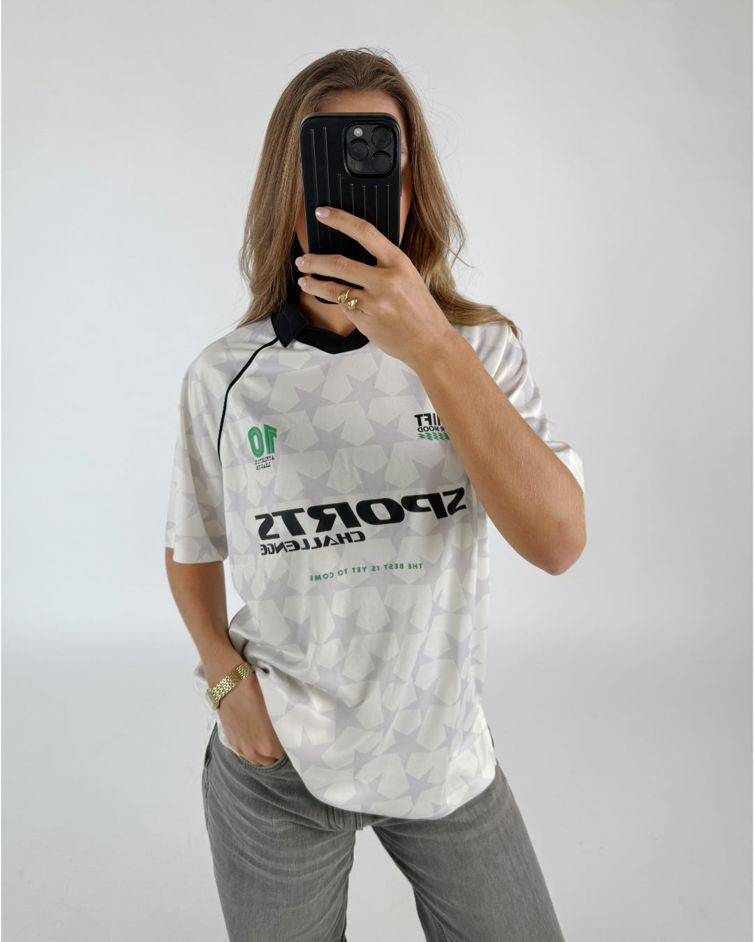 En kvinde tager et spejlbillede. Hun er iført en hvid trøje med stjernemønster og teksten "SPORTS CHALLENGE. THE BEST IS YET TO COME." Baggrunden er hvid.