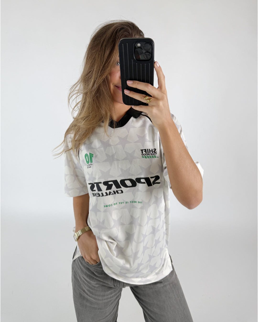 En kvinde tager et selfie med sin telefon. Hun bærer en hvid trøje med stjerner og teksten "SPORTS CHALLENGE THE BEST IS YET TO COME" og "SHIFT YOUR MOOD". Baggrunden er hvid.
