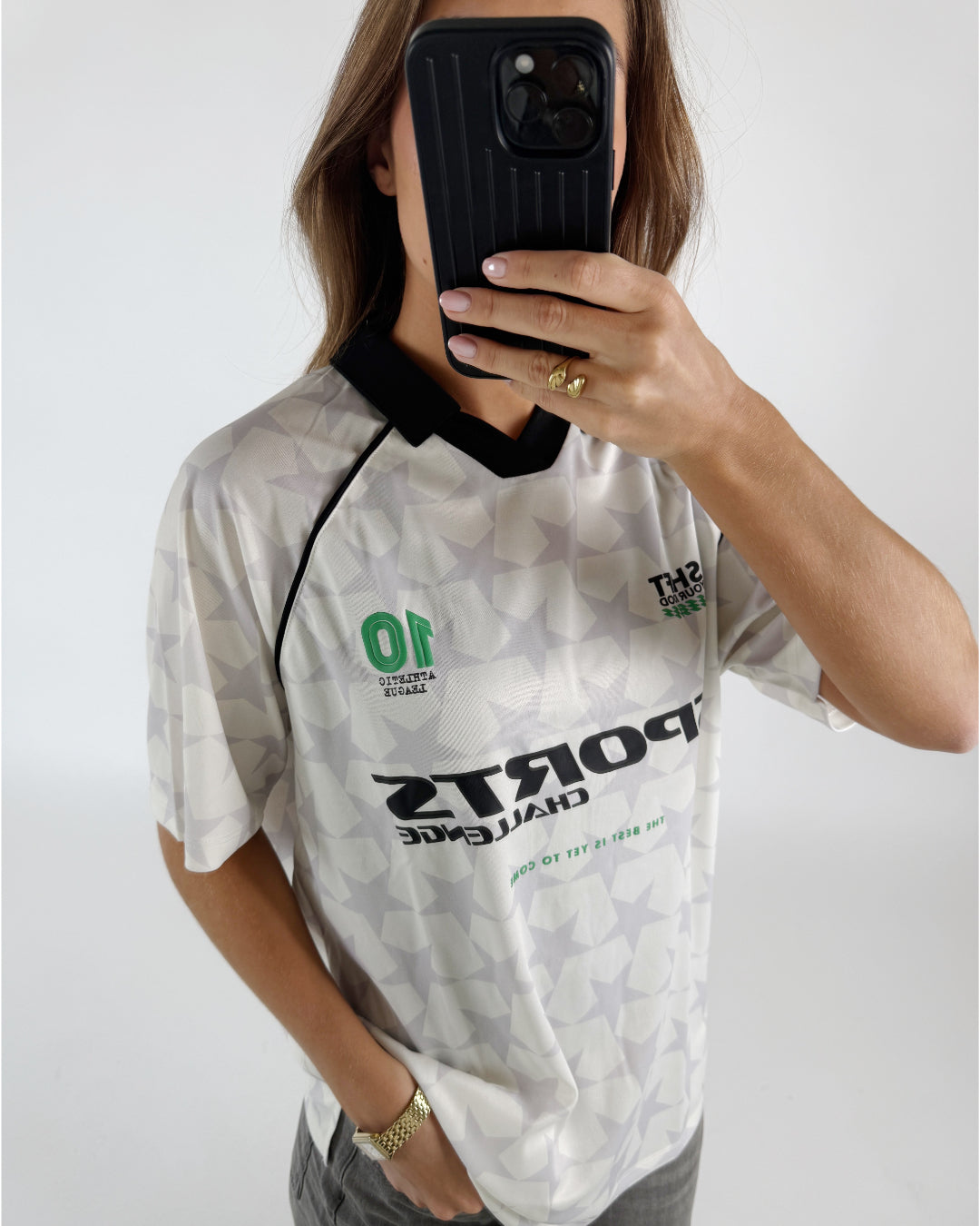 En kvinde tager et billede af sig selv med en telefon foran ansigtet. Hun er iført en retro fodboldtrøje med stjernemønster og teksten "SPORTS CHALLENGE THE BEST IS YET TO COME".