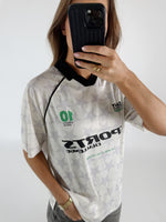 En kvinde tager et billede af sig selv med en telefon foran ansigtet. Hun er iført en retro fodboldtrøje med stjernemønster og teksten "SPORTS CHALLENGE THE BEST IS YET TO COME".