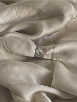 En sølvring samler lyse silkestofbølger i en blød, afslappet foldning. Stoffet skaber en glat og elegant tekstur i en neutral setting.