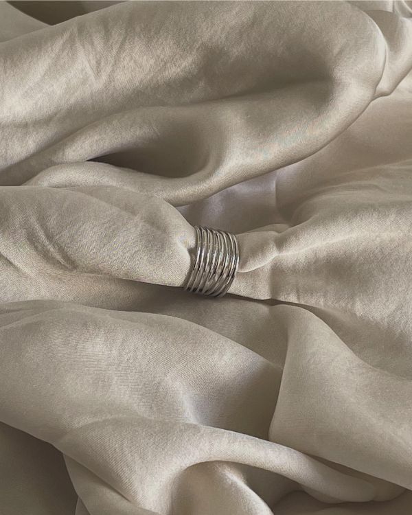 En sølvring samler lyse silkestofbølger i en blød, afslappet foldning. Stoffet skaber en glat og elegant tekstur i en neutral setting.